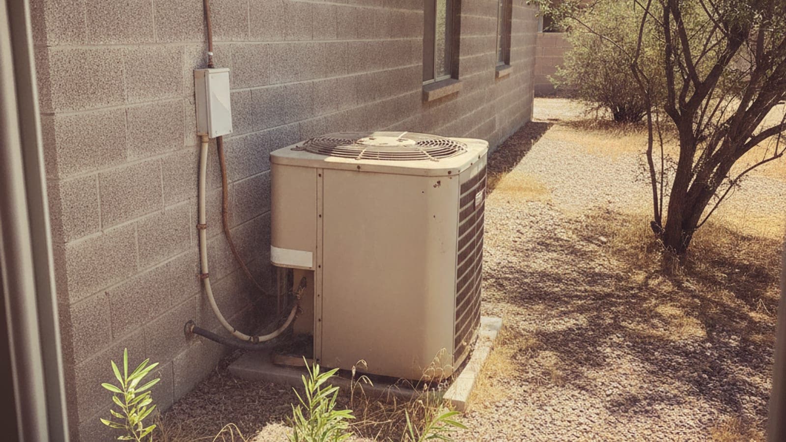 Condensador de AC viejo con la pintura descolorida junto a una casa de bloque en Tucson y un patio de grava bajo el fuerte sol del desierto